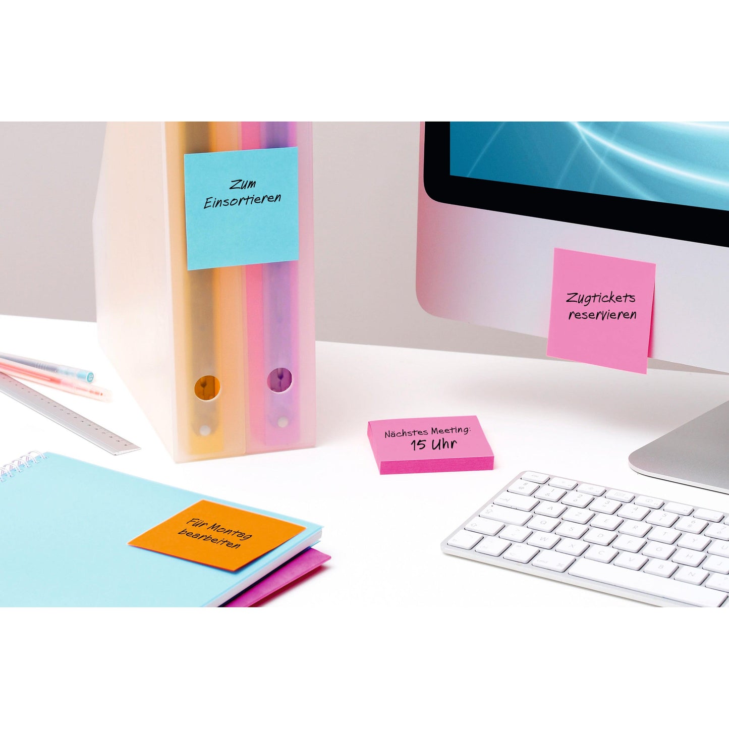 Ein Schreibtisch mit pastellfarbenen Ordnern, einem geschlossenen blauen Notizbuch, einer Tastatur, einem iMac und mehreren 3M Deutschland GmbH Post-it® Super Sticky Notes Gelb (76 mm x 76 mm) mit handschriftlichen deutschen Erinnerungen auf Ordnern, Notizbuch, Schreibtisch und Bildschirm.