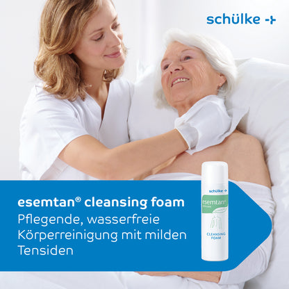 Een verzorger in een witte jas wast zachtjes de schouder van een oudere vrouw in bed. Op de voorgrond is een 500-ml-fles van het reinigingsschuim Esemtan® van Schülke & Mayr GmbH te zien, waarop de zachte reiniging van de huid wordt beschreven.