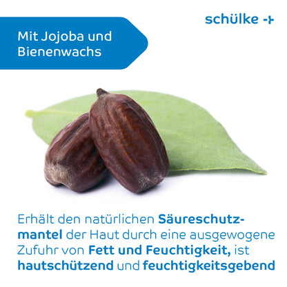 Afgebeeld is een close-up van twee bruine zaden die op een groen blad rusten. Op de achtergrond is een blauwe vorm met de tekst 'Met jojoba en bijenwas' te zien. Daaronder legt een Duitse tekst de huidbeschermende en vochtinbrengende voordelen van deze ingrediënten in de Schülke esemtan® huidlotion van Schülke & Mayr GmbH uit.